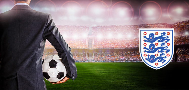 The image shows a man in a suit standing in a football stadium, holding a soccer ball under his arm. The background features a stadium filled with cheering fans under bright stadium lights. The right side of the image prominently displays the England national football team crest, featuring three lions and ten red roses.