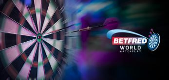 The image features a dartboard in the background with a dart in mid-flight, appearing to have been thrown towards the board. The left side of the image showcases the dartboard, with the numbers and segments clearly visible, while the dart is captured in sharp focus against a blurred background, emphasising motion and action. On the right side, the logo for the Betfred World Matchplay is prominently displayed, including the Betfred branding and a stylised dartboard graphic. The overall composition suggests dynamic movement and excitement, highlighting the competitive nature of the World Matchplay Darts event.