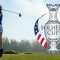 A golfer in action on a lush green golf course, swinging a club with the Solheim Cup logo prominently displayed on the right, featuring the American and European flags.