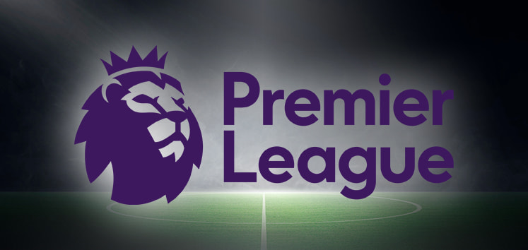 The image shows the Premier League logo, featuring a stylized lion's head wearing a crown on the left, with the text "Premier League" written in bold purple letters next to it. The background depicts a floodlit football pitch, suggesting a night match setting. The overall theme conveys the official branding of the English Premier League, the top-tier football competition in England.