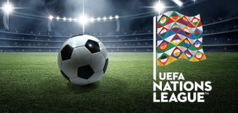 The image shows a football on a green pitch inside a stadium with bright floodlights illuminating the scene. On the right side, there is a colorful, geometric flag associated with the UEFA Nations League, accompanied by the tournament's official logo and text reading "UEFA Nations League." The stadium is empty, and the focus is on the football and the Nations League branding, symbolizing the competition.