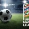 The image shows a football on a green pitch inside a stadium with bright floodlights illuminating the scene. On the right side, there is a colorful, geometric flag associated with the UEFA Nations League, accompanied by the tournament's official logo and text reading "UEFA Nations League." The stadium is empty, and the focus is on the football and the Nations League branding, symbolizing the competition.