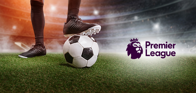 A football player wearing black boots and socks stands poised with one foot on a football on a vibrant green pitch, set against a blurred stadium backdrop. The Premier League logo is prominently displayed to the right, symbolising the competitive nature of the league.