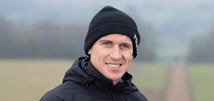 Paddy Brennan wearing a black beanie and a dark jacket stands outdoors on a path with a blurred background of fields and trees.